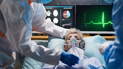 En patient med iltmaske ligger i en hospitalsseng, overvåget af sundhedspersonale.